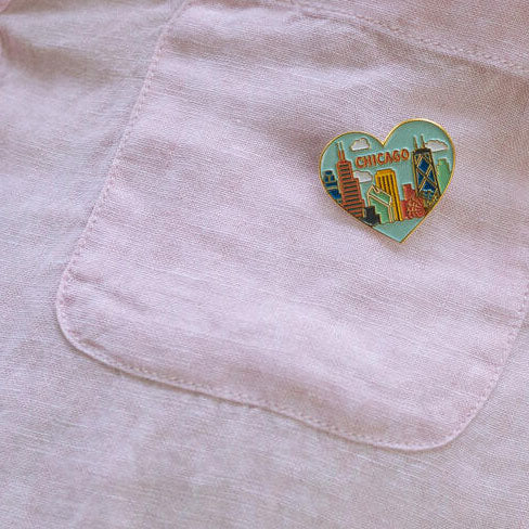A heart-shaped enamel pin with a colorful Chicago skyline design is attached to the pocket of a light pink shirt.