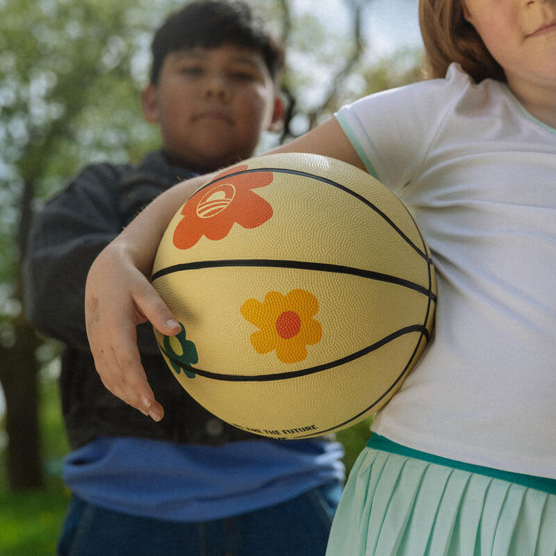 Two children are outdoors; one holds a cream-colored basketball decorated with colorful flower designs. The child in front wears a white shirt and teal skirt. The background is blurred greenery.