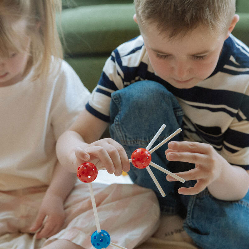 Two young children sit on the floor building a structure using colorful connecting sticks and balls, focused on assembling the pieces together.