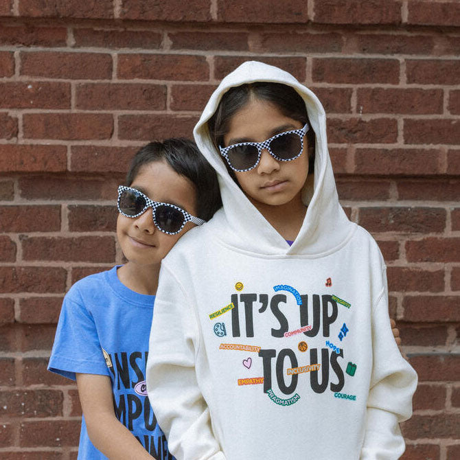 Two children wearing sunglasses stand in front of a brick wall. One child in a white hoodie with Its Up To Us on it; the other wears a blue shirt and hugs the first child from behind with a smile.