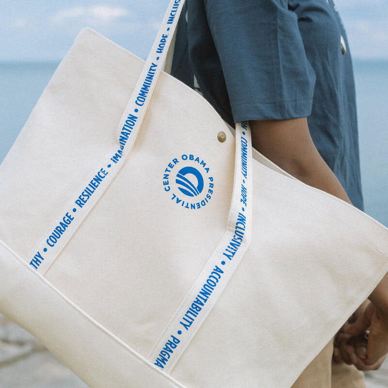 A person in a light denim shirt holds a cream-colored tote bag with blue logo and striped handles in a cozy cafe with plants and wooden tables.