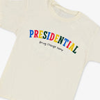 The Presidential Bring Change Home Youth T-Shirt is an off-white tee featuring PRESIDENTIAL in bold, colorful letters with Bring Change Home below in black—celebrating the Obama Foundation’s mission for positive change.