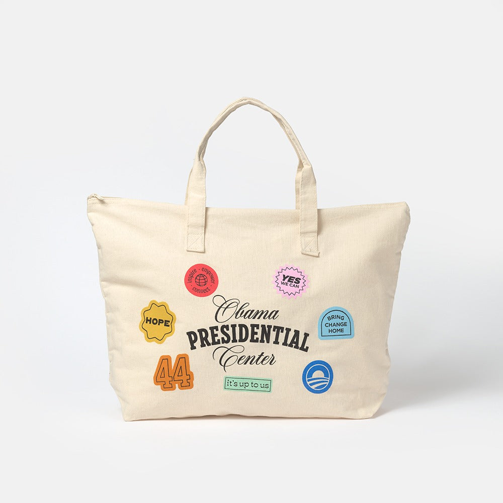 Obama Presidential Center Stickers Tote—beige canvas tote with two handles, decorated on the front with colorful patches, "44," "HOPE," badges, and slogans from the Obama Foundation collection.