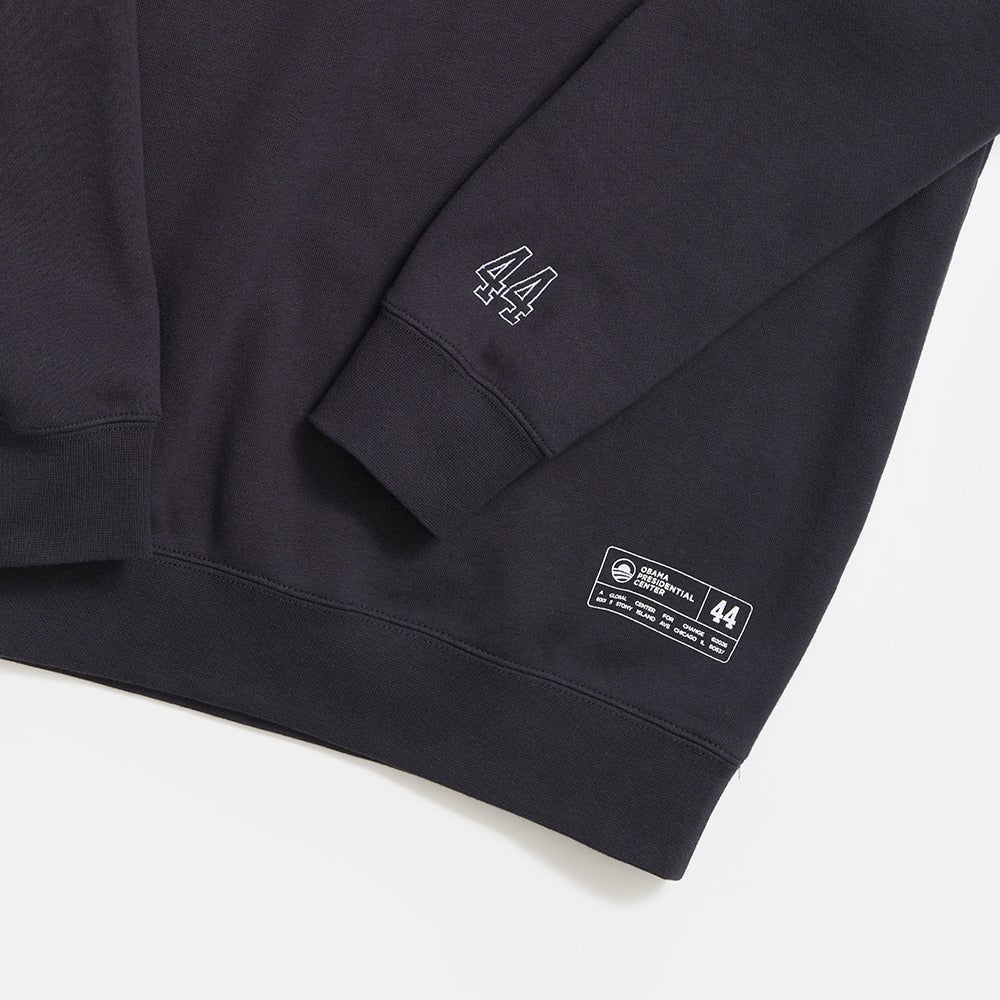 A close-up of the Obama Presidential Center Chicago Script Sweatshirt shows the number 44 stitched on the sleeve and a label with text and numbers, including 44, near the hem. The dark sweatshirt is set against a white background.