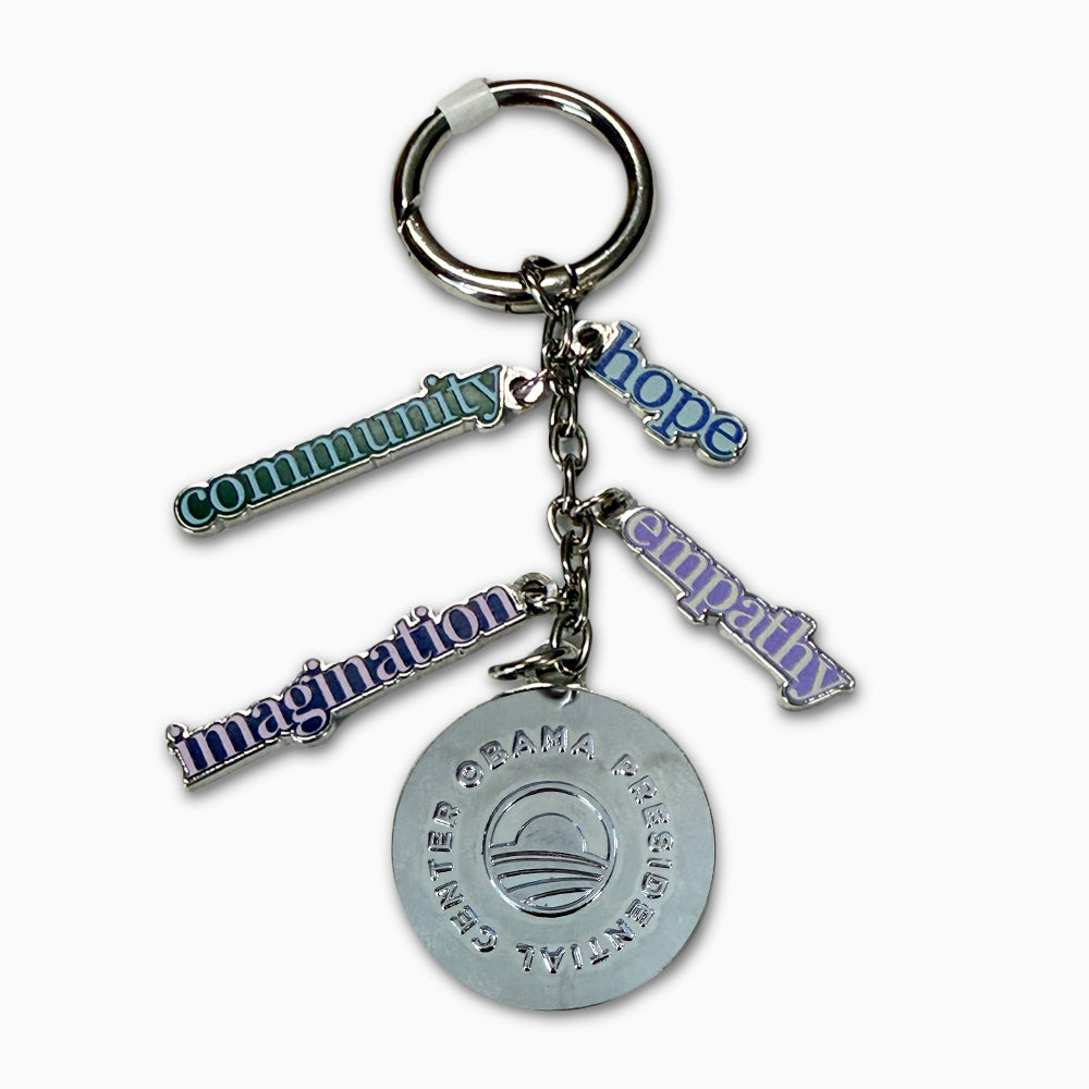 The Obama Presidential Center Dangle Keychain features four charms—green community, blue hope, purple imagination, silver empathy—and a round silver charm engraved with the Center's name and stylized logo.