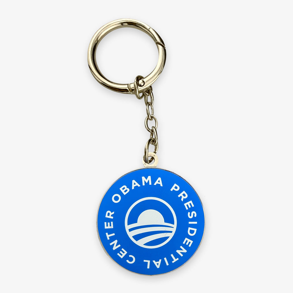 The Obama Presidential Center Logo Keychain is a round blue keychain featuring a white circular logo and "OBAMA PRESIDENTIAL CENTER" text, celebrating the Foundation’s mission in Chicago, with a metal key ring and chain.