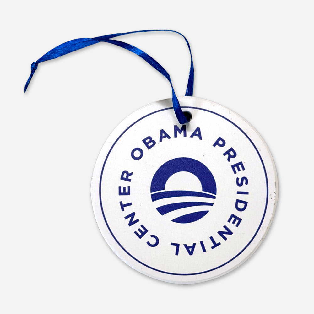 The Obama Presidential Center Logo Ornament is a round white ornament with a blue ribbon, featuring the center's logo and text in a circle, celebrating the Obama Foundation's South Side Chicago roots.