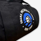 The Obama Presidential Center Home Court Duffel Bag features black cotton canvas with a basketball and blue arcs logo—an official Obama Foundation item.