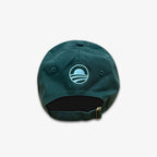 The Obama Presidential Center Community Cap is dark green, shown from the back with an adjustable strap and a light blue embroidered circular logo of curved sunrise or sunset lines—an official cap.
