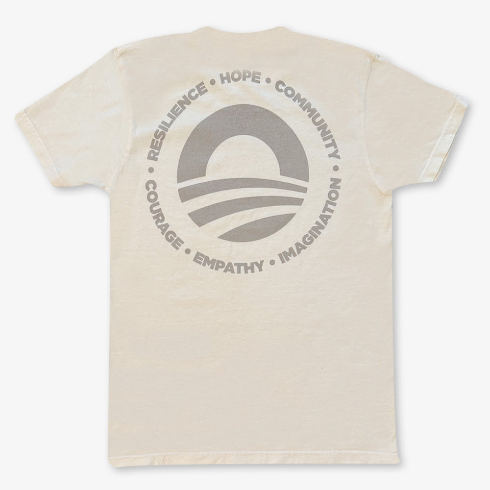 The Obama Presidential Center Hope to Action T-Shirt – Ivory features a large circular logo on the back with a rising sun, stripes, and words like RESILIENCE, HOPE, and COMMUNITY inspired by the Obama Foundation.