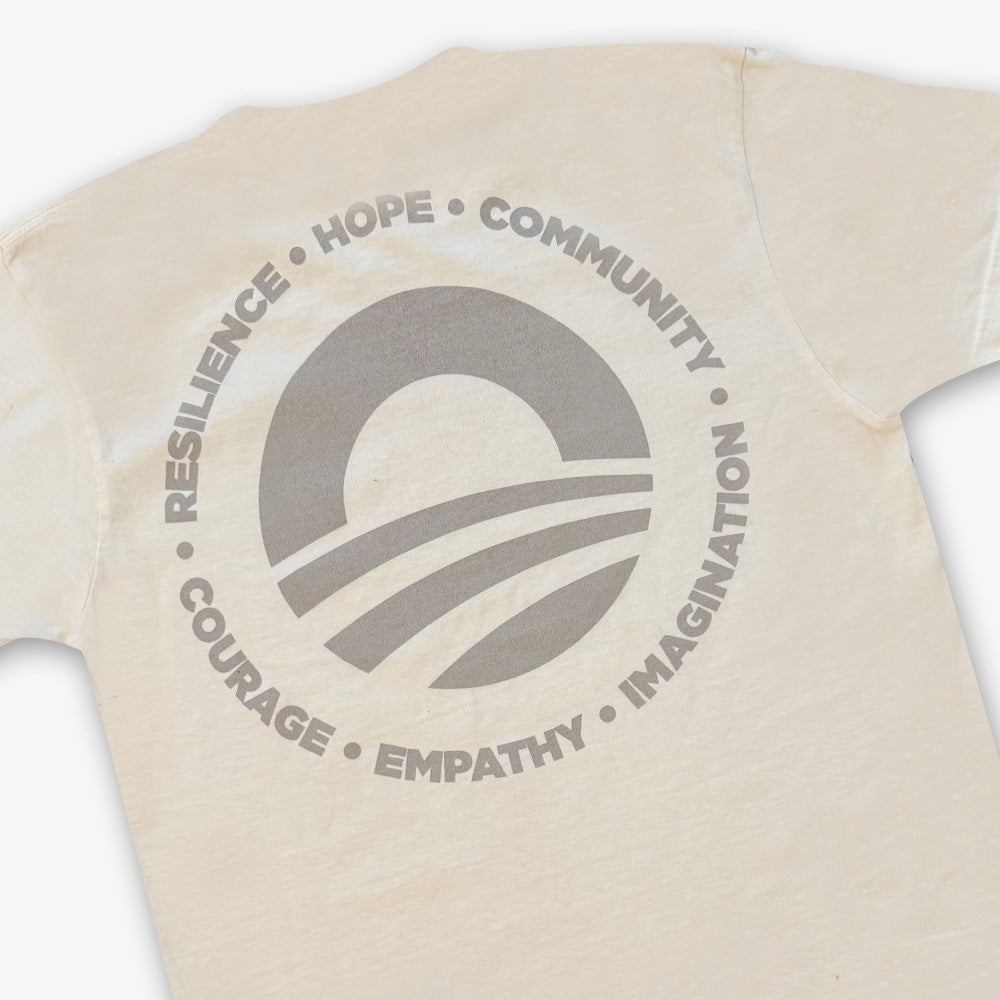 The Obama Presidential Center Hope to Action T-Shirt in ivory features a large circular sunrise logo on the back with the words Resilience, Hope, Community, Imagination, Empathy, and Courage.