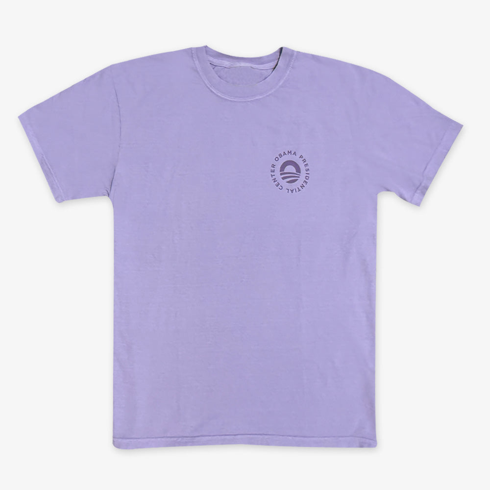 The Obama Presidential Center Hope to Action T-shirt in light purple features a small circular logo with a rising sun and “Obama Presidential Center” text on the left chest, shown laid flat on a white background.