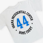 The Obama Presidential Center Home Court T-shirt is white with "OBAMA PRESIDENTIAL CENTER HOME COURT" in black lettering on the front and a bold blue number 44 printed on the back.