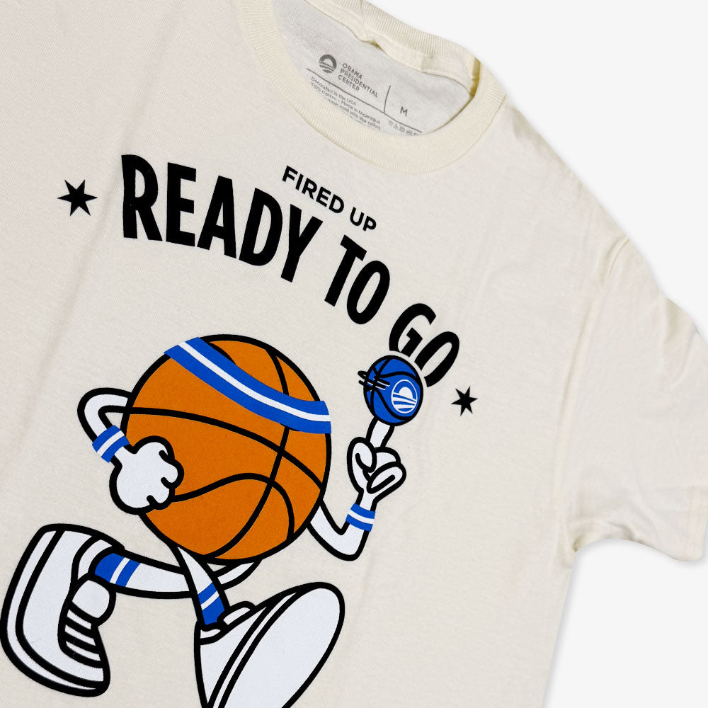 The Obama Presidential Center Home Court Youth T-Shirt is cream-colored and features a cartoon basketball character spinning a ball, bold “FIRED UP READY TO GO” text above, and small stars on each side.