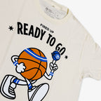 The Obama Presidential Center Home Court Youth T-Shirt is cream-colored and features a cartoon basketball character spinning a ball, bold “FIRED UP READY TO GO” text above, and small stars on each side.