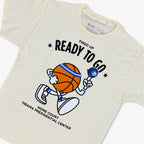 The Obama Presidential Center Home Court Youth T-Shirt is cream-colored and features a cartoon basketball holding a microphone with the phrases “Fired Up Ready To Go” and “Home Court Obama Presidential Center.”.