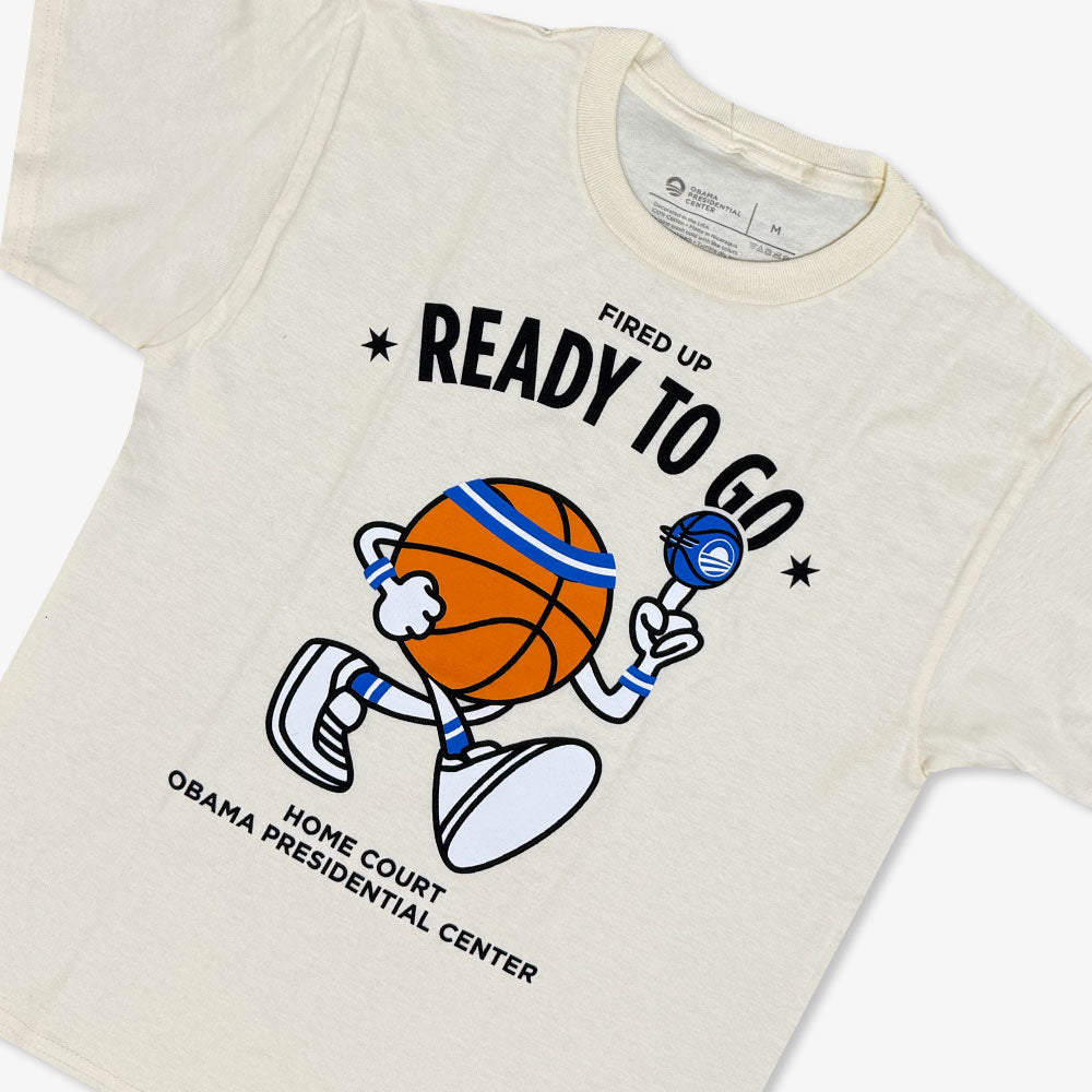 The Obama Presidential Center Home Court Youth T-Shirt is cream-colored and features a cartoon basketball holding a microphone with the phrases “Fired Up Ready To Go” and “Home Court Obama Presidential Center.”.