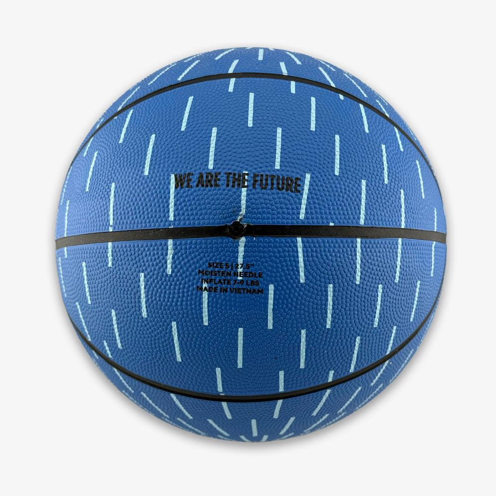 Introducing the Obama Presidential Center Blue Basketball—a kids-size basketball in a striking blue shade with light blue vertical dash patterns, black seams, and "WE ARE THE FUTURE" printed above sizing and manufacturing details. This exclusive piece is part of the Home Court collection for the Obama Presidential Center.