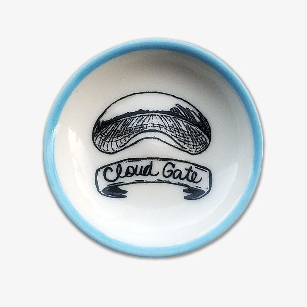 The Chicago Bean Trinket Dish is a handmade white porcelain souvenir featuring a black line drawing of the Chicago Bean, a light blue rim, and a ribbon with “Cloud Gate” below the illustration.