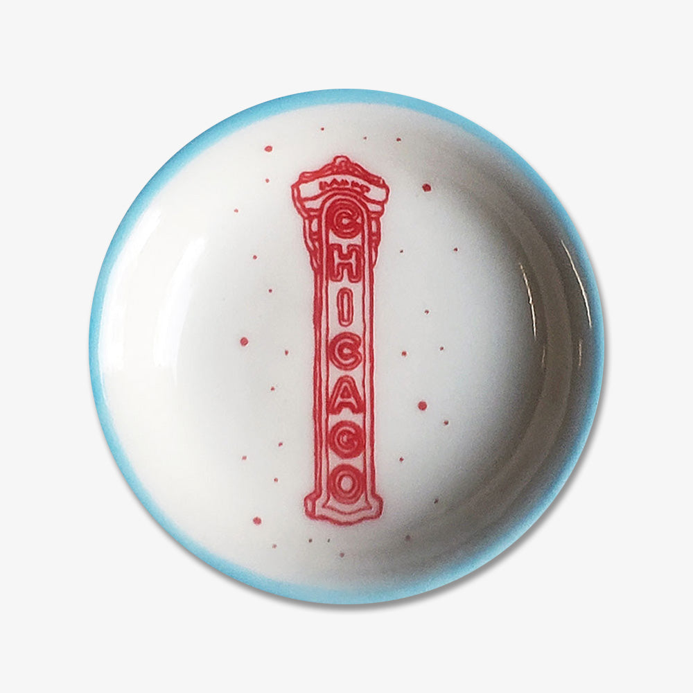The Chicago Sign Trinket Dish is a handmade round white porcelain dish with a light blue rim, red speckles, and an illustration of the iconic vertical red Chicago Theatre sign at the center.
