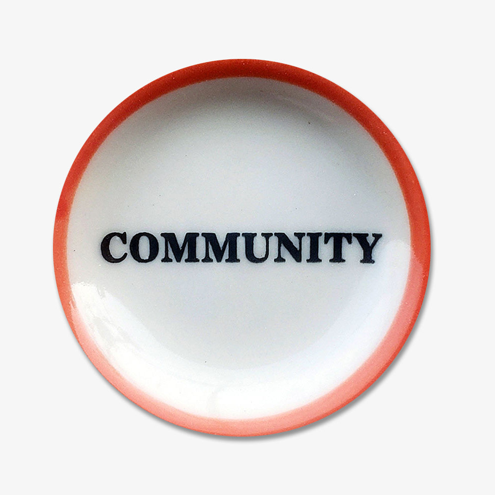 The Community Trinket Dish is a round, white handmade porcelain plate featuring a red rim and the word COMMUNITY in bold black letters at the center, inspired by the Obama Foundation.