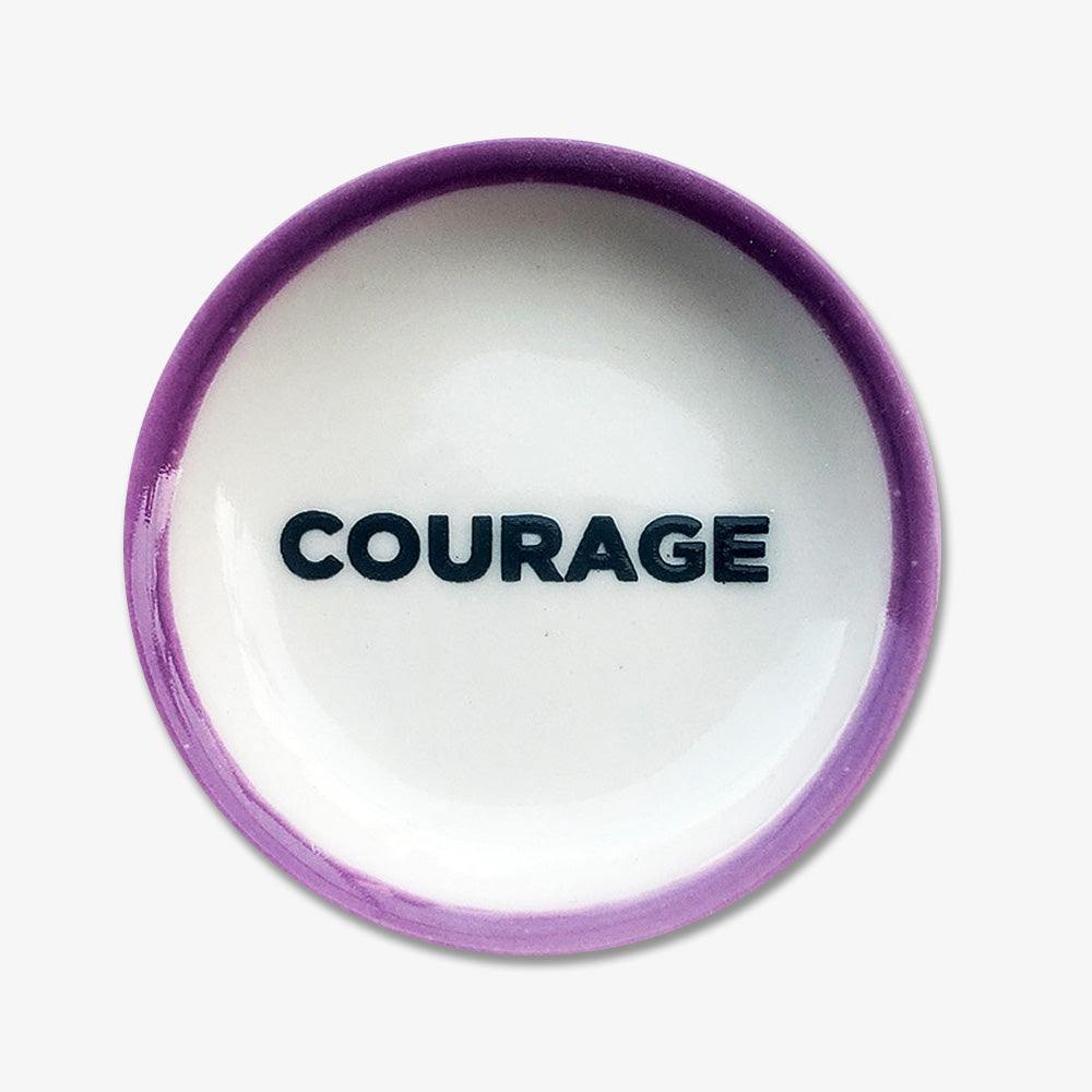 The Courage Trinket Dish is a handmade, small round porcelain dish with a purple rim and "COURAGE" in bold black letters at the center, inspired by the Obama Foundation.