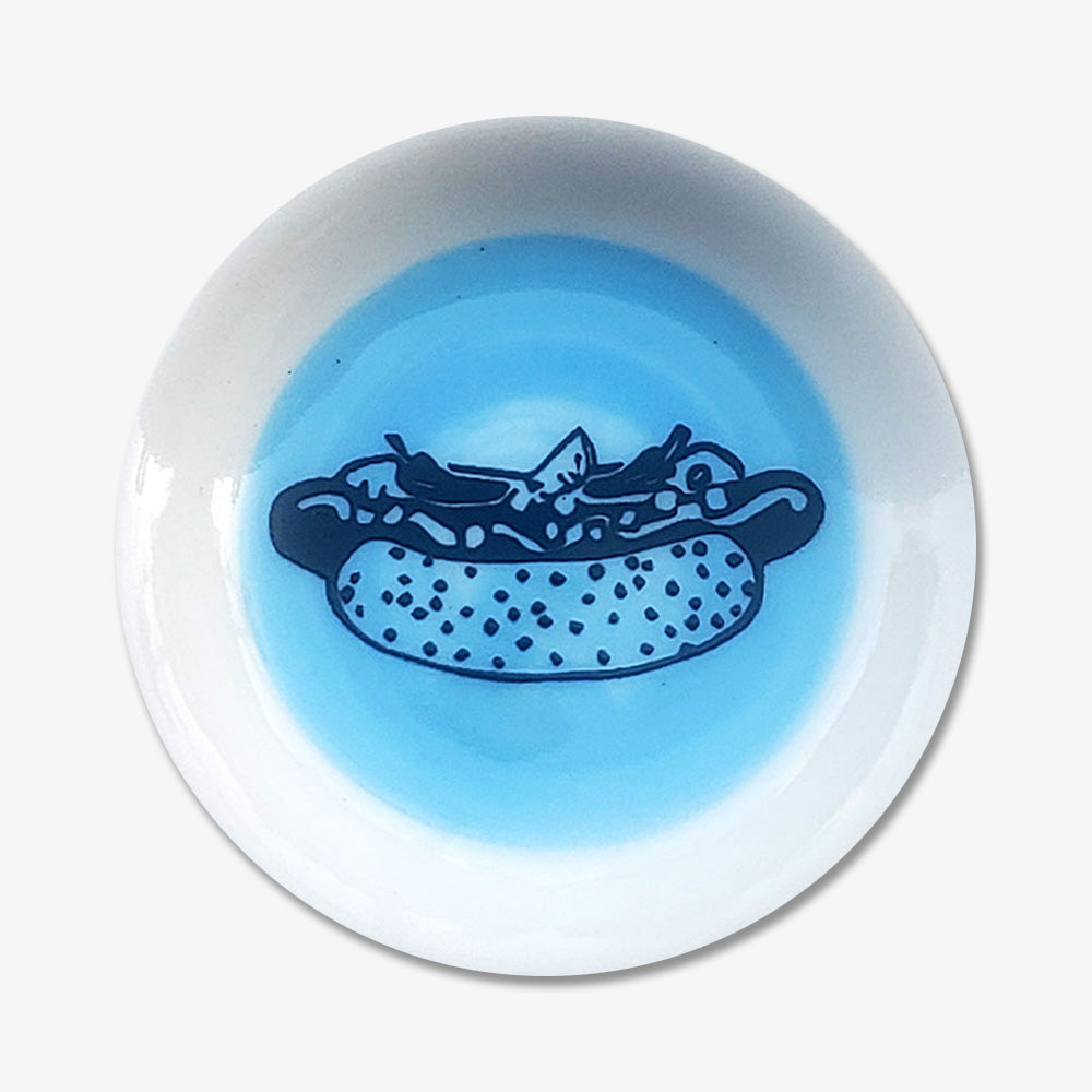 The Hot Dog Trinket Dish is a small handmade porcelain plate with a blue center, featuring an illustration of a Chicago hot dog complete with relish, mustard, and peppers—perfect for showing your love of Chicago style.
