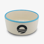 The Obama Presidential Center Pet Bowl is a white ceramic bowl with a light blue rim and black stylized sunrise logo, made by a Black-owned ceramic studio, shown against a plain white background.