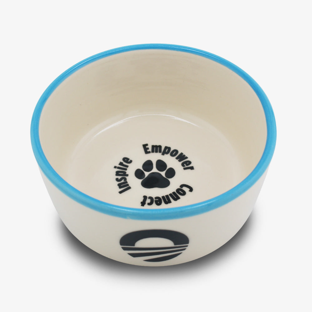 The Obama Presidential Center Pet Bowl is a ceramic pet bowl with a blue rim, handmade by a Black-owned studio. It features a black paw print and the words Inspire, Empower, and Connect inside, plus a white exterior with black logo.