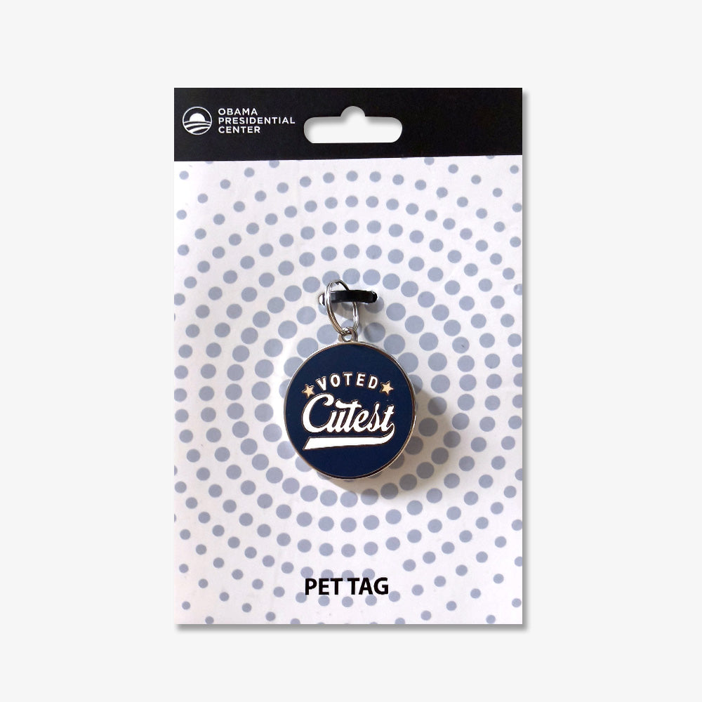 The Voted Cutest Dog Tag is a round, navy tag with “Voted Cutest” in white and gold script, attached to an Obama Presidential Center and Pet Tag card featuring a dotted circular background pattern.