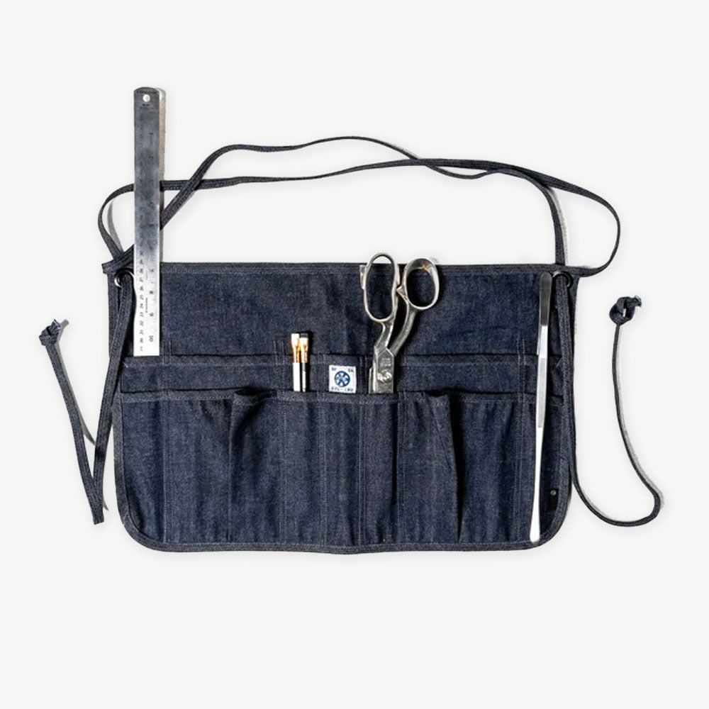 The Waist Apron is hand-crafted from 100% dark denim cotton with waist ties and a white label on the pocket. Multiple pockets hold a ruler, pencils, pen, and scissors, making this kitchen accessory stand out against a plain white background.