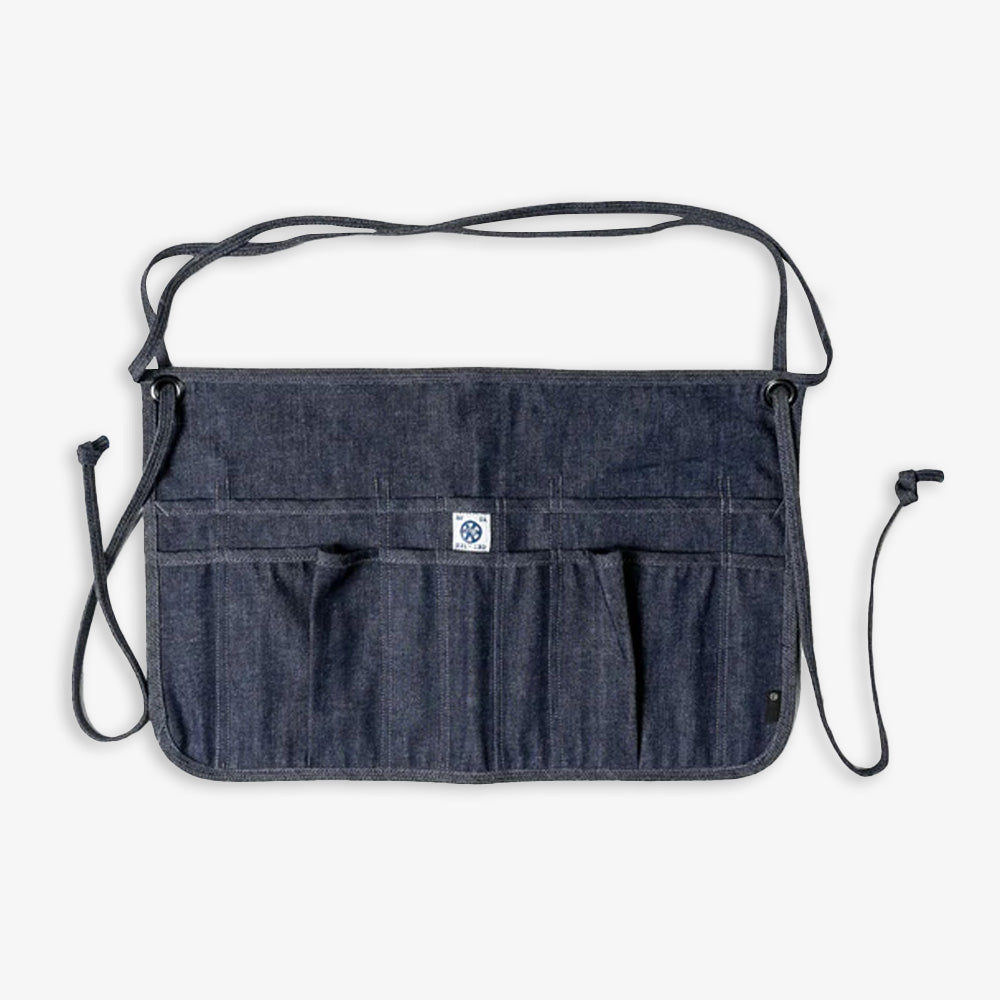 The Waist Apron, made from 100% cotton dark denim, features multiple front pockets and long adjustable straps, shown laid flat on a light background.