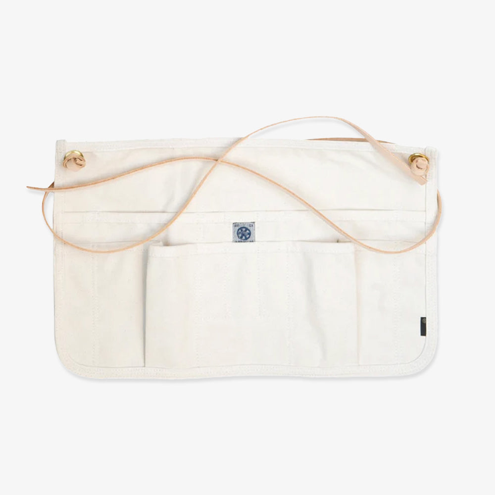 The Waist Apron is a hand-crafted kitchen accessory made from white 100% cotton, featuring three front pockets and tan leather straps, displayed flat against a white background.