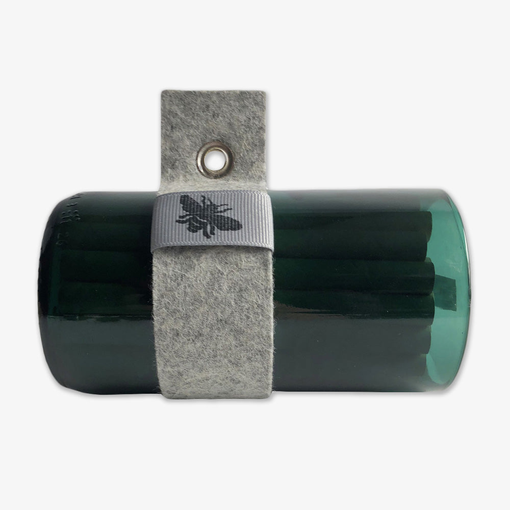 The Bee Habitat is a dark green upcycled wine bottle with a gray felt strap, metal grommet, and ribbon adorned with a black bee illustration, all set against a plain white background.