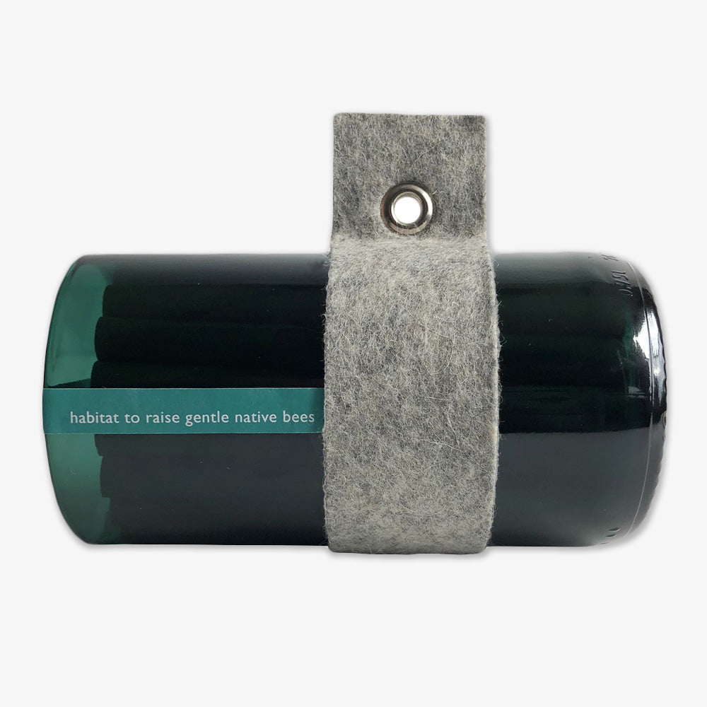 The Bee Habitat is a cylindrical, dark green upcycled wine bottle with a grey felt hanging band and metal eyelet—labeled for raising native bees. Stylish and functional, it's perfect as a native bee or mason bee house.