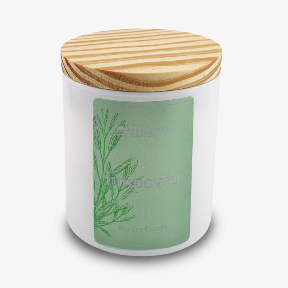 A white hand-poured candle jar with a light wooden lid and a green label reading Empower Sprig Candle 9 oz., featuring a botanical illustration on the left side of the label.