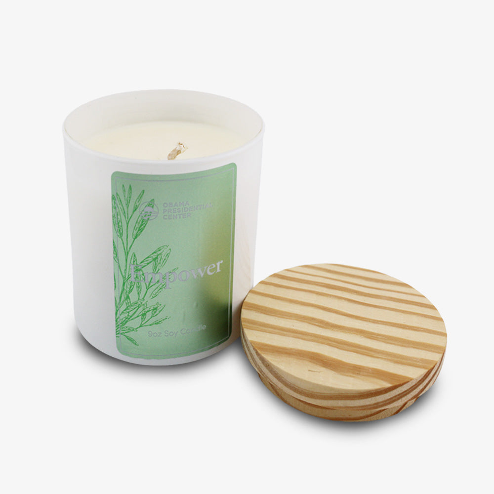 The Empower Sprig Candle 9 oz. is a hand-poured candle in a glass jar with a green botanical label and round wooden lid, placed on a white background. Your purchase supports the Obama Foundation’s mission.