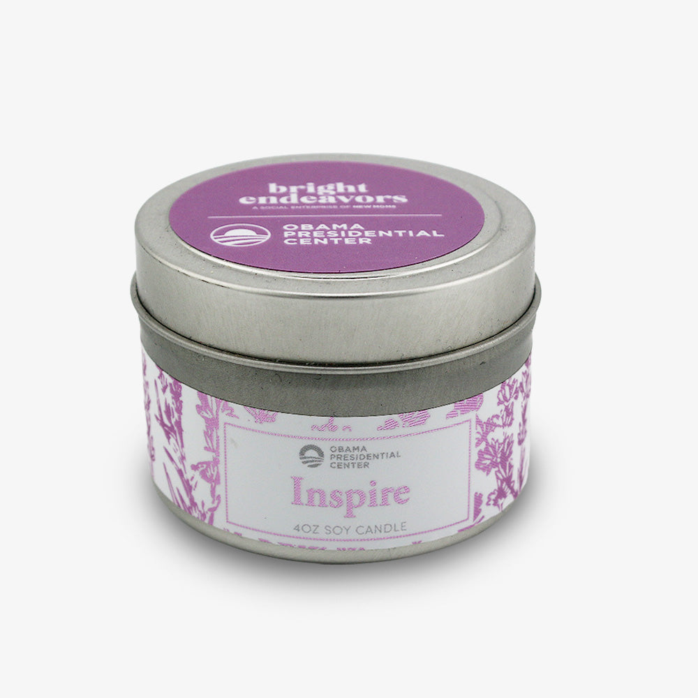 The Inspire Sprig Candle 4 oz. is a hand-poured lavender candle in a small silver tin. Its lid features a purple sticker with "bright endeavors" and Obama Foundation logos, and the tin showcases a purple floral pattern.