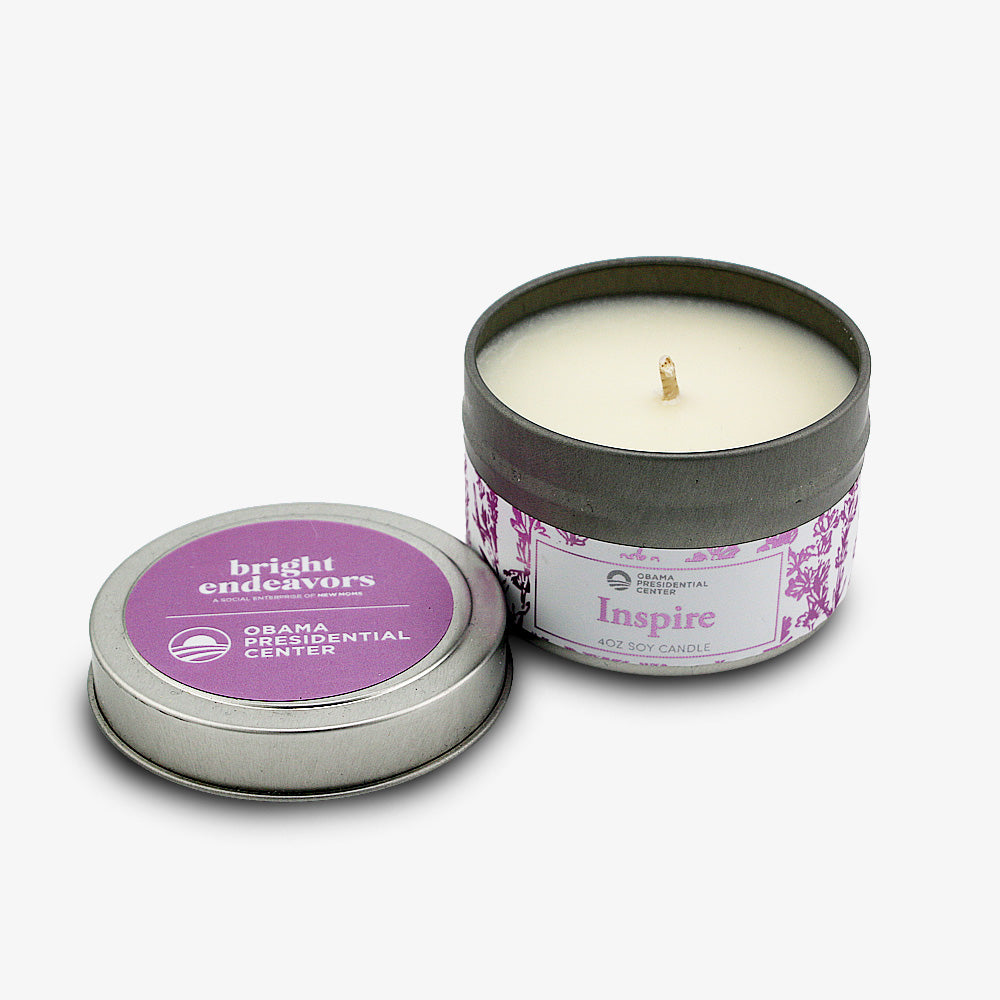 The 4 oz. Inspire Sprig Candle sits open next to its matching lid reading “bright endeavors ORNAMENTAL CENTER.” This unused, hand-poured white candle features a purple floral design on the tin.