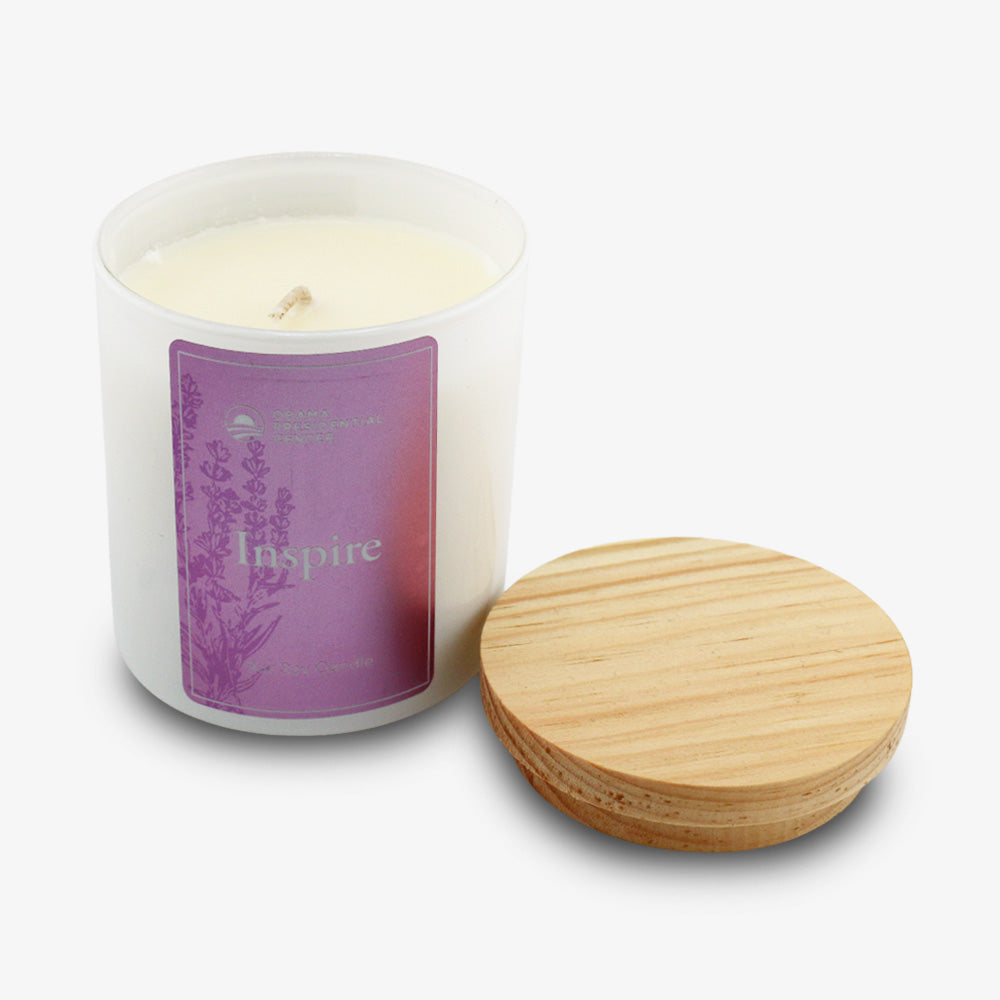 The Inspire Sprig Candle 9 oz. features hand-poured lavender wax in a frosted glass jar labeled "Inspire" with a purple floral design, accompanied by a round wooden lid, set against a white background.