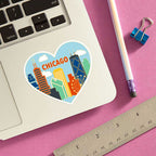 The Chicago Heart Sticker, featuring vibrant city landmarks in a heart shape, decorates a silver laptop on a pink surface with a pencil, blue binder clip, and metal ruler nearby.