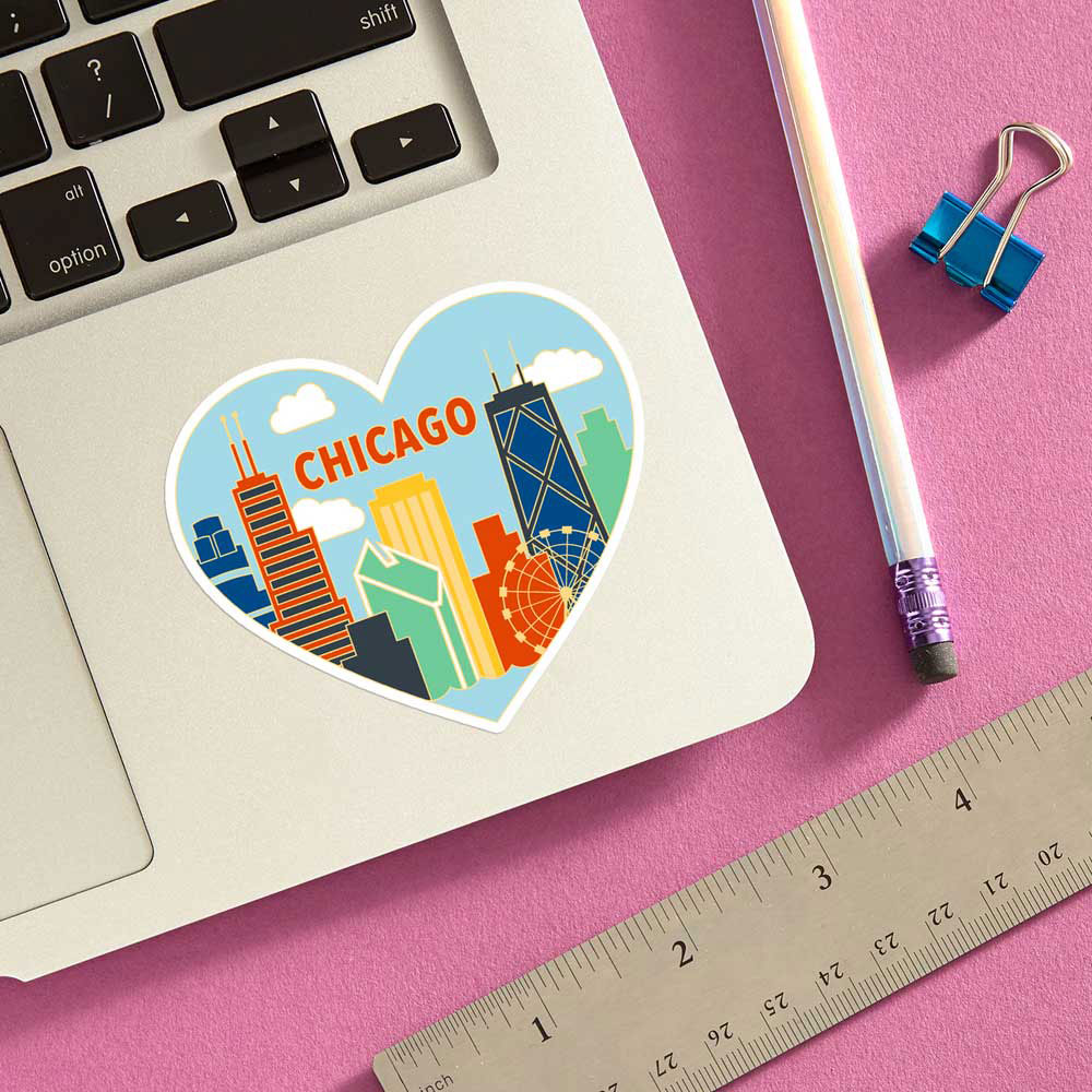 The Chicago Heart Sticker, featuring vibrant city landmarks in a heart shape, decorates a silver laptop on a pink surface with a pencil, blue binder clip, and metal ruler nearby.