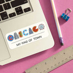 The Chicago Icons Sticker spells CHICAGO in city-themed icons with “MY KIND OF TOWN” below, shown on a laptop beside a pencil, paperclip, and ruler on pink—ideal for anyone wanting a unique Chicago souvenir.