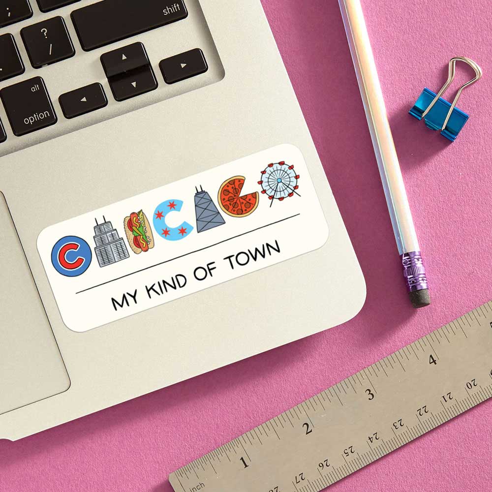 The Chicago Icons Sticker spells CHICAGO in city-themed icons with “MY KIND OF TOWN” below, shown on a laptop beside a pencil, paperclip, and ruler on pink—ideal for anyone wanting a unique Chicago souvenir.