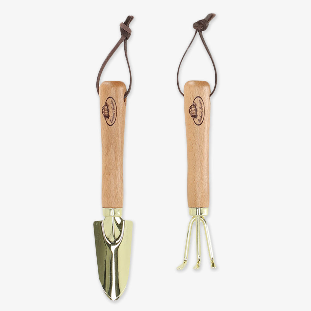 The Shovel & Rake Mini Garden Tools include a beech wood-handled trowel and three-pronged cultivator, both with shiny stainless steel ends, leather loops, and a logo on the handle.