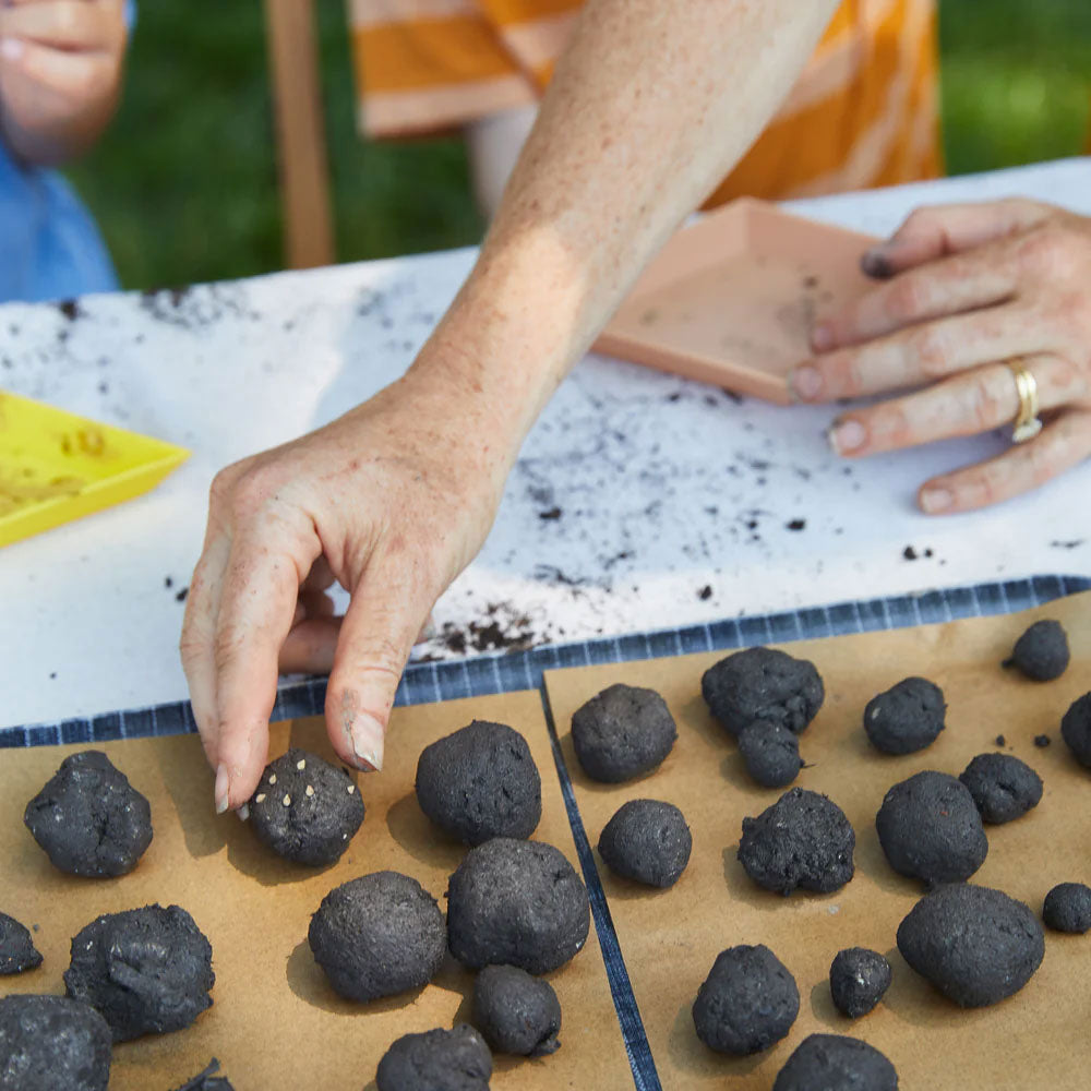 A person places a small black ball from the Pizza Garden Seed Ball Kit onto brown paper with other seed balls. Another person's hands and scattered dirt on the table suggest sustainable gardening in progress.