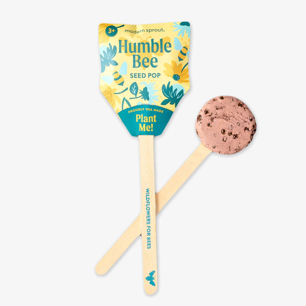 A lollipop-shaped Pollinator Seed Pop with “Plant Me!” instructions sits beside an unwrapped one showing its soil-like, seed-filled texture—perfect for sustainable gardening and attracting pollinators.