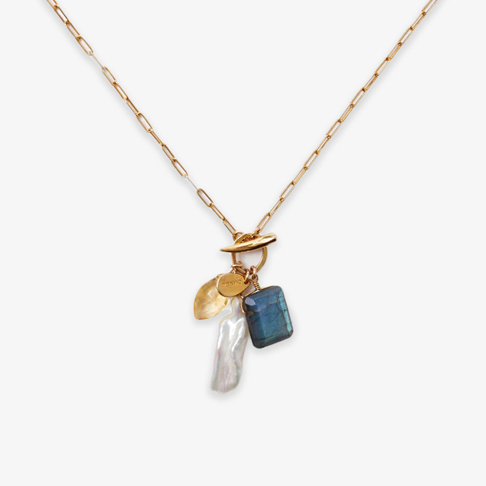 The Gemstone Mix Charm Necklace is a gold paperclip chain with a toggle clasp, featuring four charms—a gold disc, yellow gem, blue gemstone, and white pearl—crafted in sustainable artisan jewelry style with travel-inspired details.