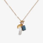 The Gemstone Mix Charm Necklace is a gold paperclip chain with a toggle clasp, featuring four charms—a gold disc, yellow gem, blue gemstone, and white pearl—crafted in sustainable artisan jewelry style with travel-inspired details.