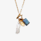 The Gemstone Mix Charm Necklace features a gold paperclip chain with three pendants—a white pearl, yellow gemstone, and blue rectangular stone—crafted as sustainable artisan jewelry.